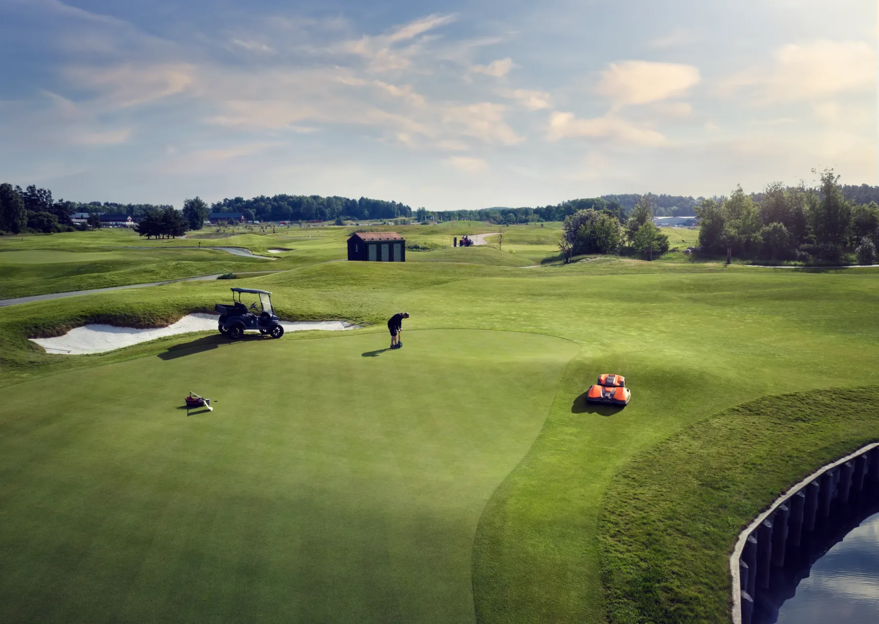 TP-126157. En person puttar på en välskött golfgreen omgiven av sandfällor, med en golfbil och två gräsmatteskötselmaskiner i närheten under en delvis molnig himmel.